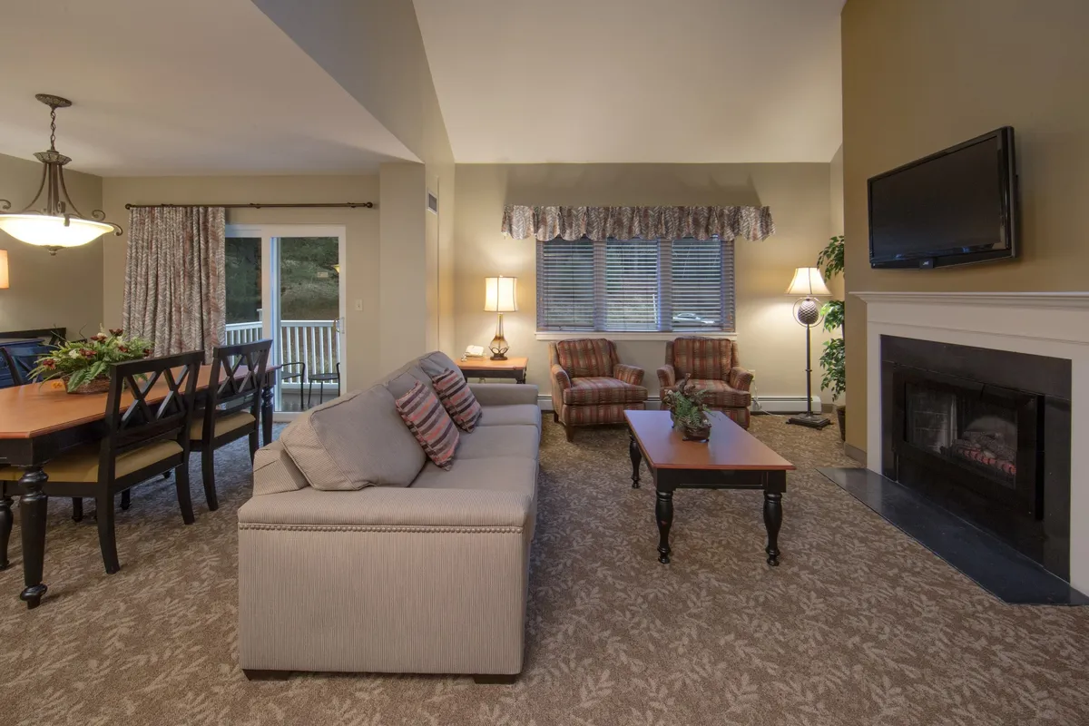 The image showcases a warm, inviting open-plan living area with a plush sofa positioned beside a glowing fireplace, a spacious dining table perfect for group meals, and large windows letting in natural light. The setting is ideal for relaxing after adventures in Vermont's outdoors, gathering for movie nights, or enjoying intimate family dinners—all with the cozy ambiance of the fire and mountain views just outside.