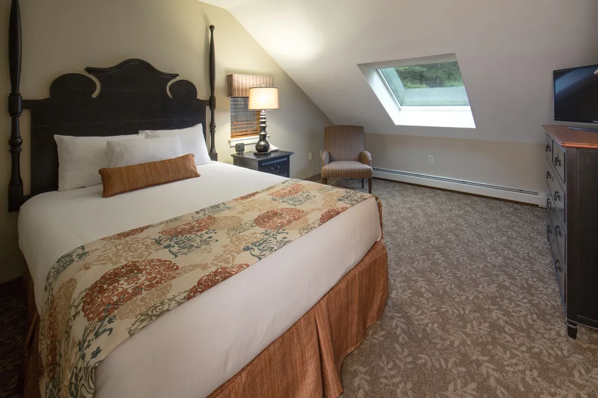 This cozy interior bedroom features a bed with an ornate headboard, decorative bedding, a sloped ceiling with a skylight that lets in natural light, a nightstand with a lamp, and a comfy chair by the window. The soft, tranquil tones and reading nook create a peaceful retreat after a long day.