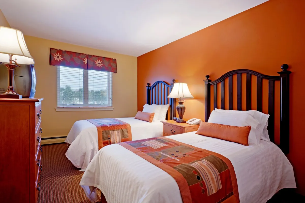 This inviting bedroom showcases two beds with decorative headboards, colorful bedding, bedside tables with lamps, and a window streaming in natural light. The layout offers ample sleeping space for families or groups, with a welcoming, homey ambiance.