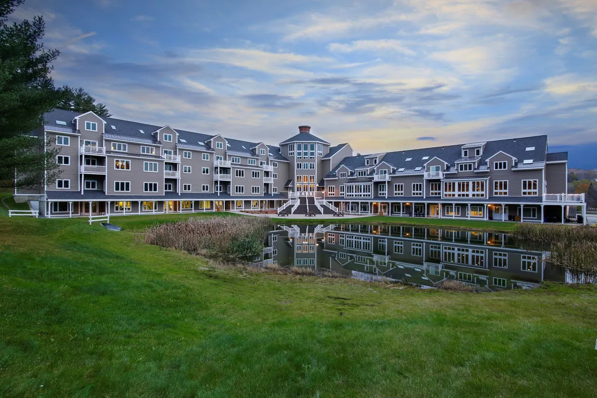 A wide view of the resort's central building reflected beautifully in calm water, bordered by grassy areas and trees. Guests are seen strolling along the pathways and relaxing on the lawns, while the light mist and reflections add a magical touch to the natural setting.