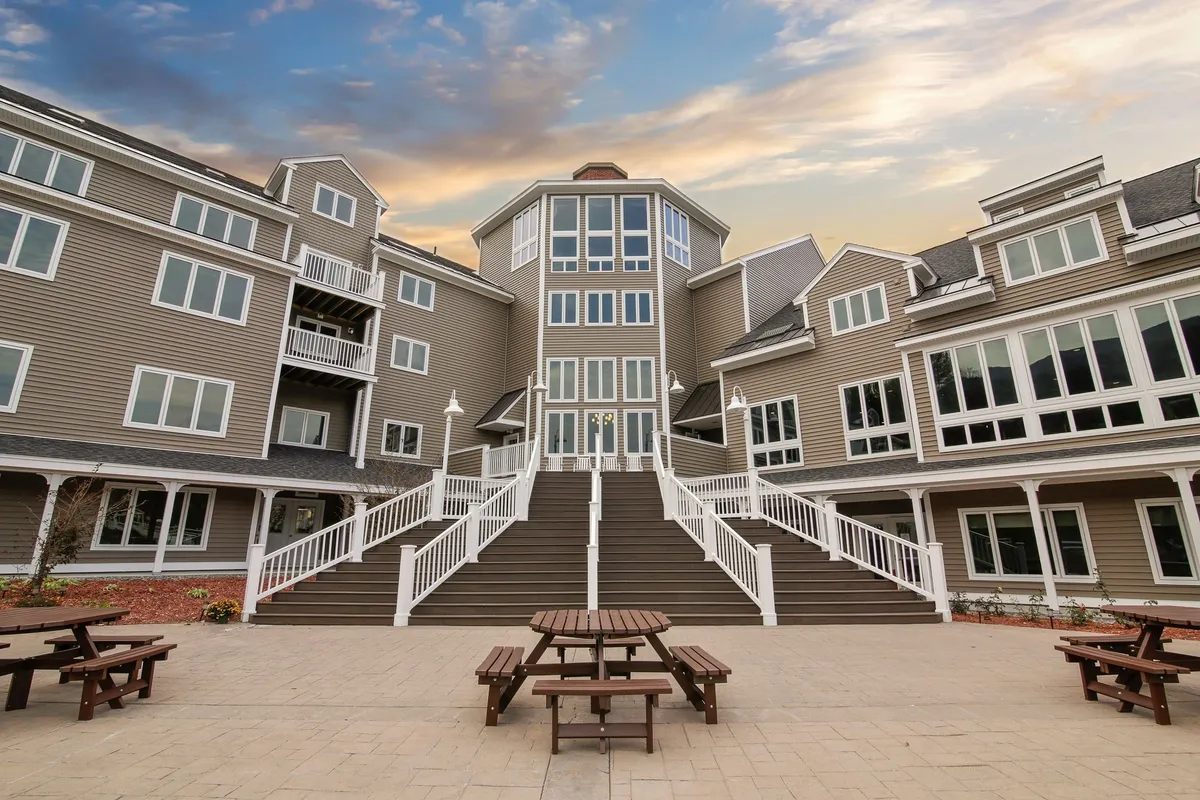 This scene captures the resort’s exterior grand staircase, large windows, and outdoor picnic tables. The mood is welcoming, with sunset lighting creating a warm ambiance. Guests are shown enjoying the patio, perhaps after a day of adventure, with community events or casual gatherings taking place outside. The image conveys the transition from outdoor activities to relaxed social time at the property.