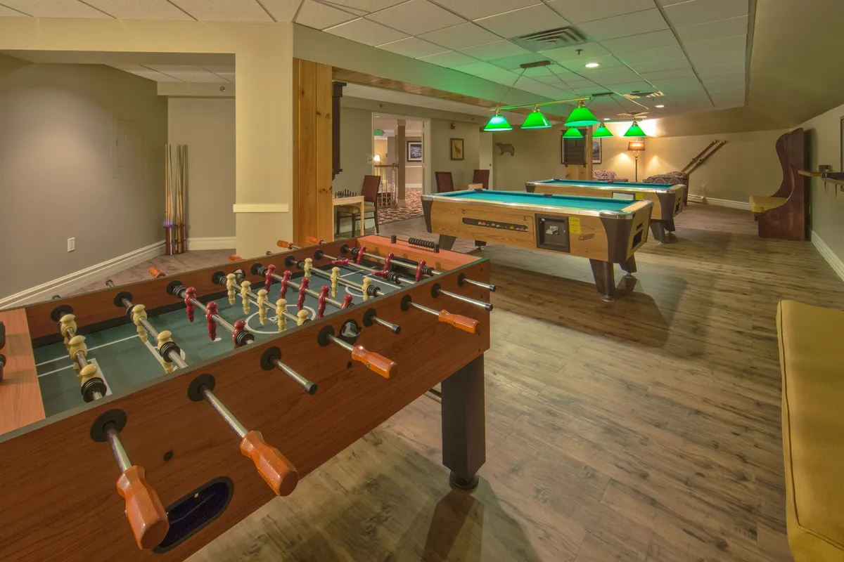 A vibrant game area featuring pool tables and a foosball table, accented by hanging green lights. Groups of friends are engaged in play, laughter, and socializing, evoking a sense of fun and community.