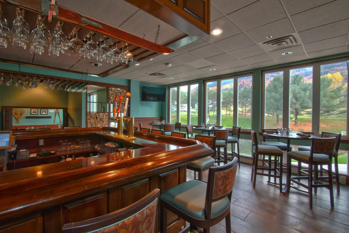 This image showcases a stylish indoor dining area featuring a bar counter, large windows allowing natural light, tables and chairs set for meals, and decorative wall art. The ambiance is relaxed and social, perfect for gathering, dining, or enjoying drinks together. The setup hints at flexible dining options, both formal and casual.
