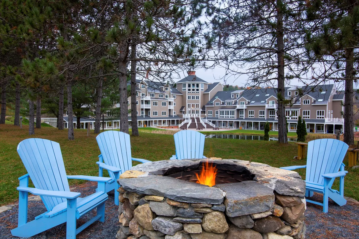 A cozy outdoor fire pit area is depicted, with blue Adirondack chairs arranged in a circle and the hotel building softly illuminated in the background. People are gathered around the fire, enjoying s'mores and relaxed conversation as the sunset colors the sky.